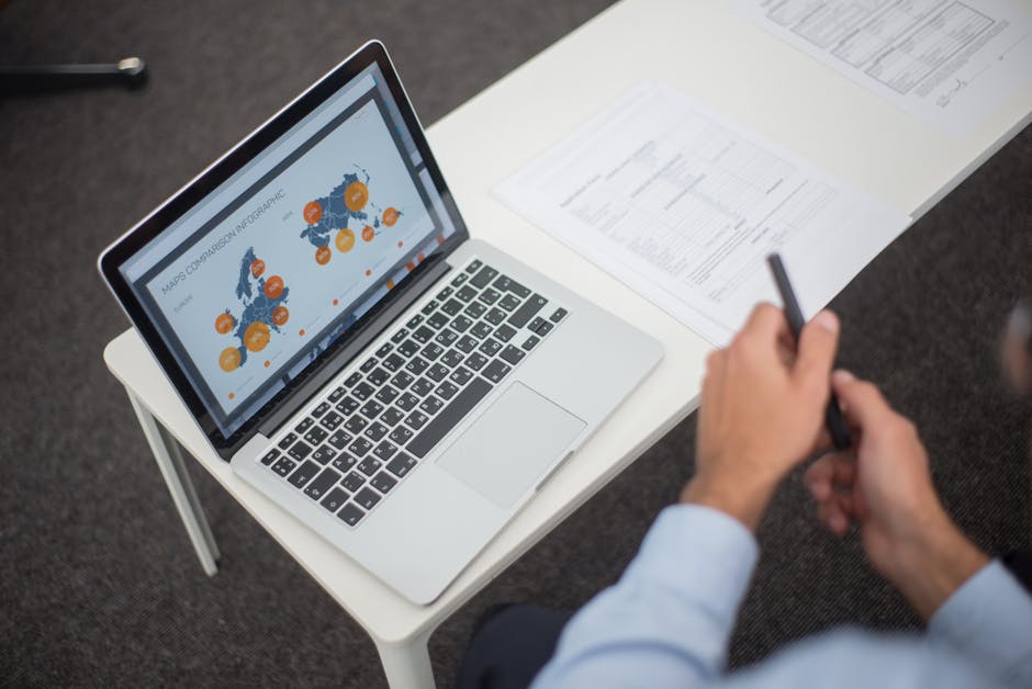 Professional analyzing data infographics on a laptop at an office desk, featuring documents and a pen.
